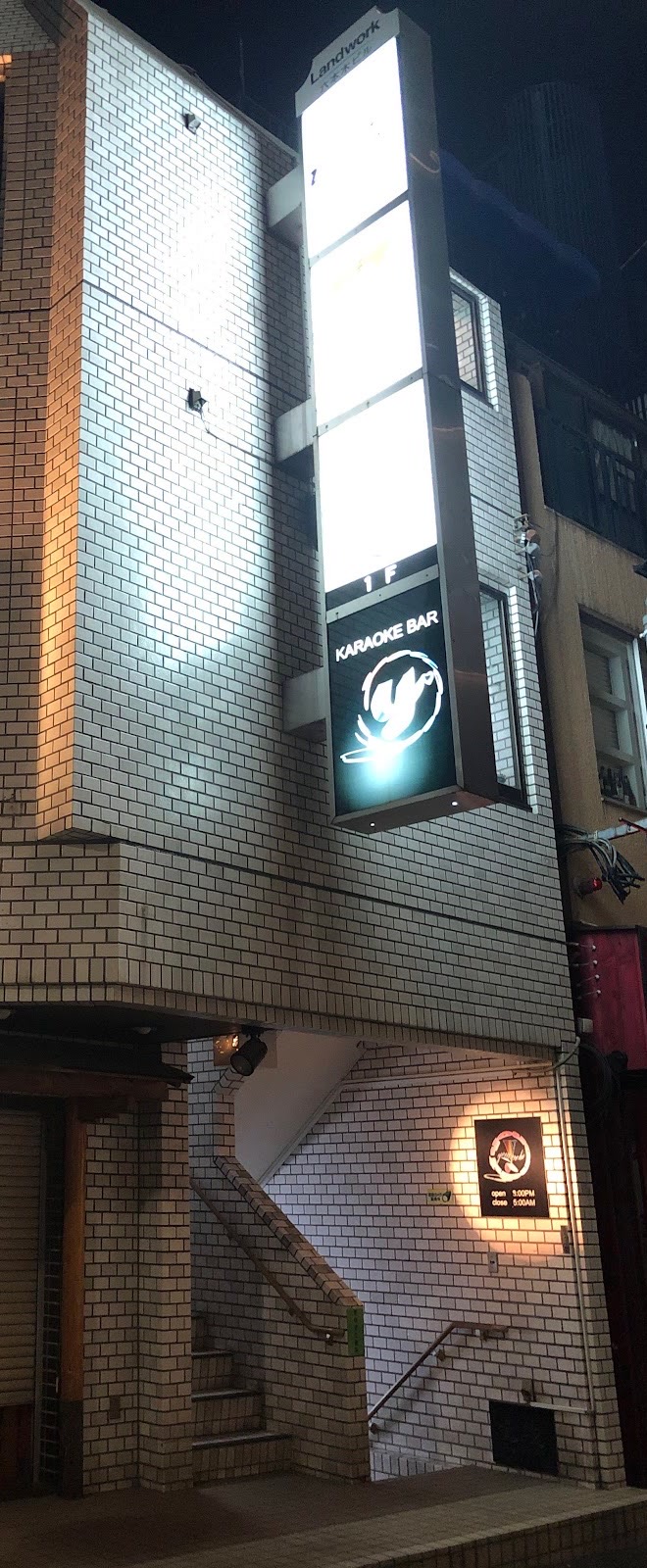夜の白いタイル張りのビル。縦長の看板塔のいちばん下に、「KARAOKE BAR」の文字と白い「y」のマークが入った黒いパネルが灯る。下の階段室の壁には「open 9:00PM close 5:00AM」と書かれた小さな黒い看板。
