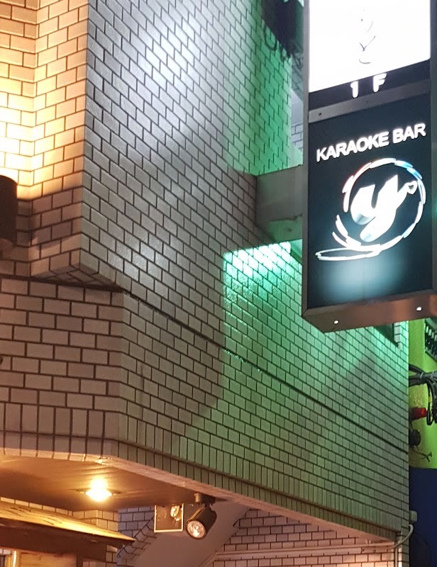 夜、緑がかった光に照らされた白いタイルの壁。看板塔の黒いパネルに「KARAOKE BAR」の文字と白い「y」のマーク。